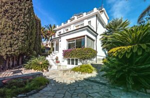 A cozy stone cottage with a with lush vegetation of flowers and a green garden surrounded by palms and trees in Liguria, Italy.